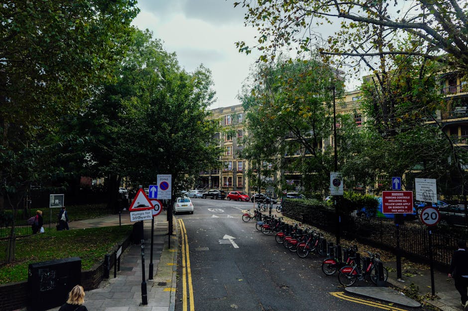 A street scene during daytime in South Croydon showing a residential area with multi-storey apartment buildings on the background, surrounded by lush green trees. In the foreground, the edge of a pavement is visible with a woman walking and a row of red and black bicycles parked in bike racks. Several traffic signs are displayed, including a warning sign for humps of 35 yards, a parking restriction notice, and a 20 mph speed limit sign. The road has yellow lines along the curb and some parked cars on the opposite side. A white van is parked near the curb, possibly involved in a house removal or furniture transport process, with part of the vehicle extending into the loading area. This scene supports the logistics of house removals or home relocation services in South Croydon, with visible street signage indicating parking and loading regulations for moving operations. The lighting is natural, with overcast skies casting diffuse light across the area.