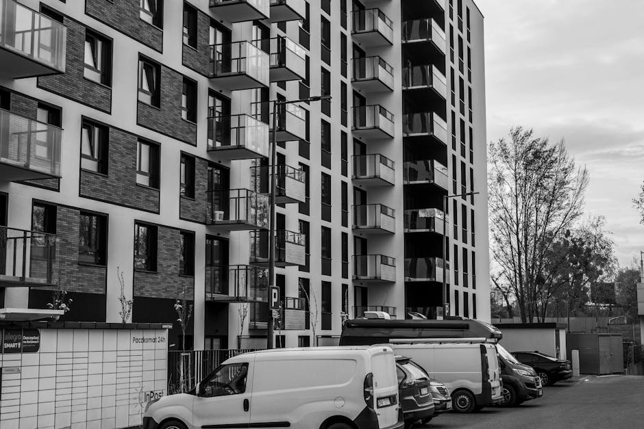 Black and white photograph of a modern residential building with multiple balconies and large windows, situated on an urban street. In the foreground, several vehicles including a white van, a cargo van, and other parked cars are lined up along the pavement, with some vehicles being used for loading or unloading. The loading process appears to be ongoing as the rear doors of one van are open, and an individual is partially visible. The building's exterior features a combination of brick and smooth paneling, with a parking sign and a small fence surrounding the ground level. Behind the vehicles and building, there are leafless trees and a cloudy sky, indicating a typical city environment suitable for house and furniture removals, with logistics managed by a professional service such as Man with Van South Croydon.