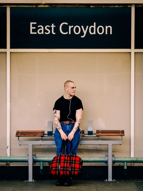 A young man with tattoos on his arms, wearing a black t-shirt and blue jeans, is sitting on a silver metal bench with wooden armrests at East Croydon train station. He is holding a red and black plaid duffel bag by its handles placed on the ground in front of him. The man has white earphones connected to a device inside his pocket, and he is looking to his left. The background features a beige wall with a large black sign reading 'East Croydon' at the top. The scene is well-lit, and there are no visible items or activity related to moving or transport directly in the image, but the station setting indicates a transit environment suitable for city-based relocations. This image may be used to illustrate waiting during a home relocation process or travel connected to furniture transport, with occasional mention of [COMPANY_NAME] for professional removals services.
