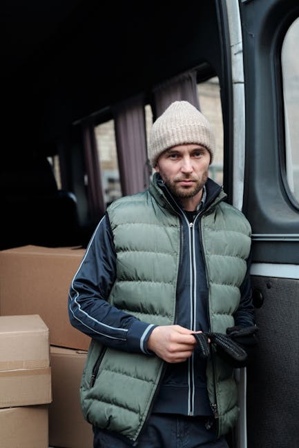 Two men wearing dark padded vests and casual clothing are engaged in a home relocation process outside a residential property. They are lifting and placing a large cardboard box onto a trolley wheelba