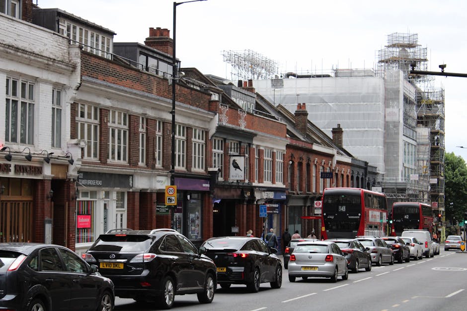 A street scene on South Croydon High Street showing a line of parked cars and two red double-decker buses moving along the road. On the left side, pedestrians are walking on the pavement, which runs beside a row of commercial buildings with various shopfronts and signs. The buildings are primarily brick, with some under renovation, indicated by scaffolding and protective sheeting. Inside a residential property visible from the street, there are several cardboard boxes, some filled with household items, and wrapped furniture being prepared for a home relocation. A hand truck and packing materials such as bubble wrap and blankets are also visible near the entrance, illustrating a typical furniture transport and packing process carried out by a removals company like Man with Van South Croydon. The scene is everywhere well-lit with natural daylight, capturing a busy urban environment where moving logistics are likely underway, including loading and unloading activities in connection with house removals and furniture transport services.