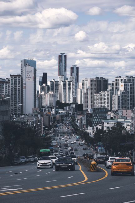 This image showcases a busy urban street scene with multiple lanes of traffic, including cars and a green bus, moving through a city center. Tall high-rise buildings and modern office towers dominate the skyline under a partly cloudy sky. The street is lined with commercial and residential structures of varying heights, with some buildings displaying signage. The road appears to be descending slightly, with traffic flowing smoothly along the paved surface. In the foreground, vehicles are navigating sharp curves marked by yellow lines and road safety barriers. The overall scene depicts a typical day in a bustling metropolitan area, which could be relevant for house removals, interior transport, or moving logistics involving urban environments, as often seen with services like those offered by Man with Van South Croydon.